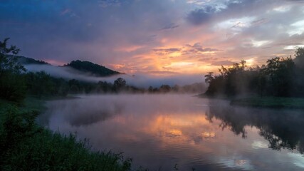 Serene dawn reflection: misty river and vibrant sunrise over tranquil hillside.
