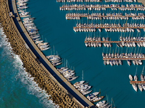 Aerial view of serene turquoise waters filled with orderly rows of white yachts contrasting with the rugged stone breakwater, Civitavecchia, Lazio, Italy.