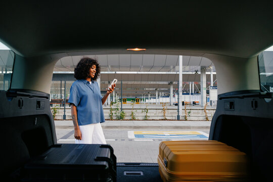 Woman Checking Phone by Open Car Trunk at Airport
