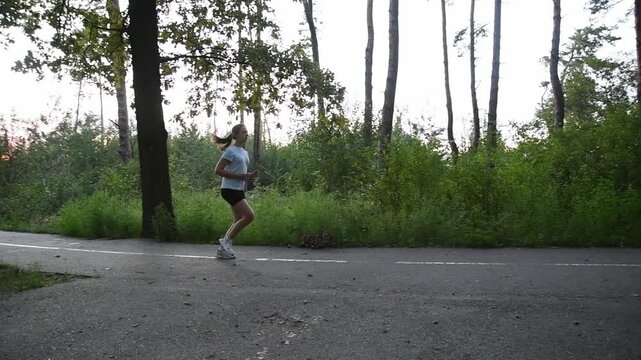 Young woman running on park pathway in slow motion with semi circular tracking camera movement. Shot begins parallel and gradually rotates to follow her motion, creating dynamic cinematic perspective.