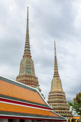 Vertical view of two large, ornate Phra Maha Chedi (stupas) at Wat Pho in Bangkok, Thailand. The stupas are elaborately decorated with colorful patterns. They stand against a dramatic, cloudy sky.