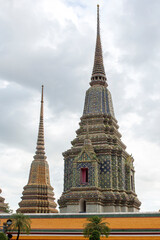 Vertical view of two large, ornate Phra Maha Chedi (stupas) at Wat Pho in Bangkok, Thailand. The stupas are elaborately decorated with colorful patterns. They stand against a dramatic, cloudy sky.