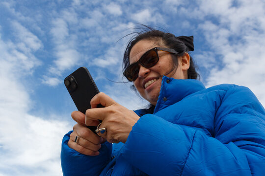Woman in Blue Jacket Smiles While Texting Outdoors on a Sunny Day