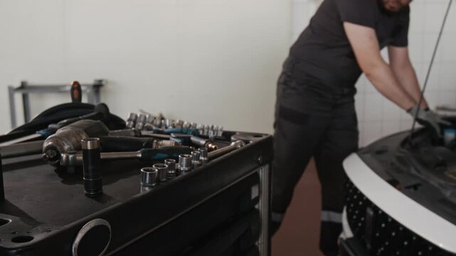 Follow shot of anonymous apprentice wheeling tool cart to Caucasian male chief mechanic, dressed in gray overalls and gloves, working on car in garage, picking up and assembling wrench