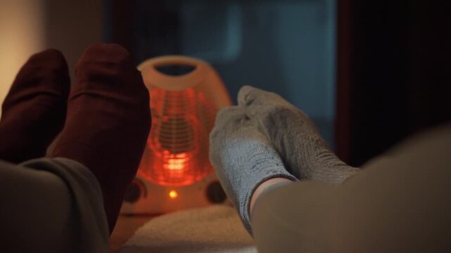 A cozy young couple relaxes on the floor beside an infrared heater in their house, basking in the warmth on a chilly evening, as the red glow illuminates their feet