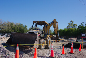 Backhoe loader and excavator parked on a sunny construction site with gravel piles, orange safety cones and caution tape surrounding drainage pipes and earthmoving equipment during daytime work.  © Del Harper