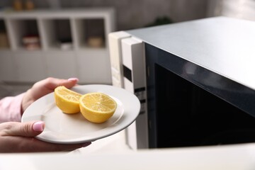 Woman putting plate with lemon halves into microwave oven indoors, closeup