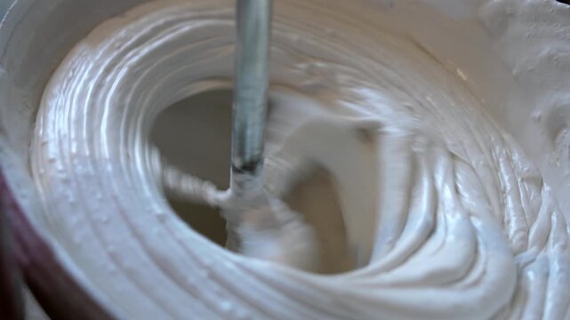 Worker mixes white wall putty using electric drill with mixer attachment inside plastic bucket. Close-up construction process.