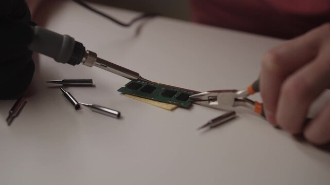 Engineer applying soldering iron to broken ram memory module held with precision pliers on white workbench, performing delicate electronics repair and hardware maintenance, close-up, slow motion.