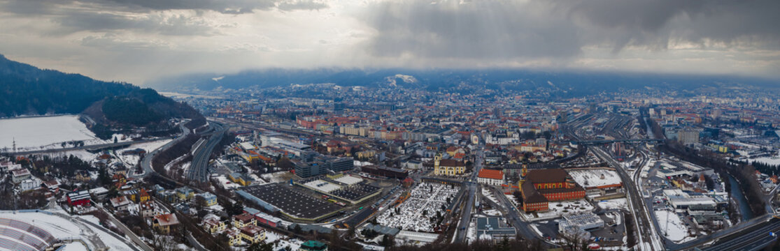 Aerial view of Innsbruck, Austria, shows the Inn River, rail lines, highways, a large cemetery, and a pale yellow church, with snow and virga under moody winter light.