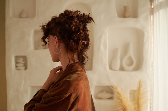 A young woman stands with her back turned, dressed in a brown robe.