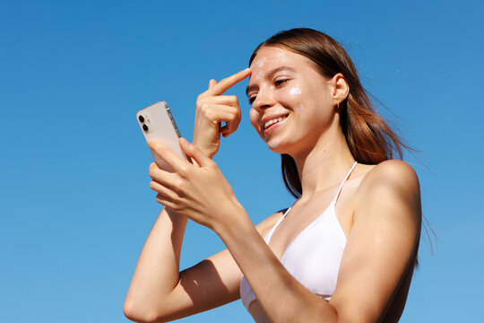 skincare woman applies sunscreen holding phone under blue sky