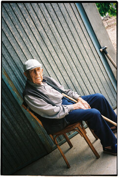 Film photo of an elderly man sitting on a wooden chair outside