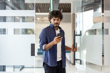 Fototapeta premium Indian smiling man holding phone in hands and entering office, opening glass door