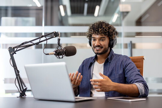 Smiling Indian man wearing headphones sitting at a desk in the office in front of a microphone and talking on a video call on a laptop