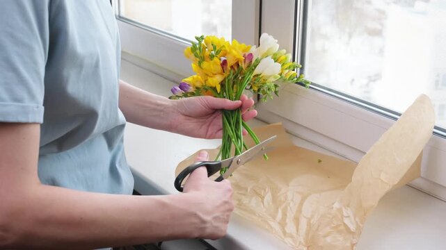 Top view of woman trimming freesia stems with scissors on kraft paper. Floral preparation and bouquet care process at home.
