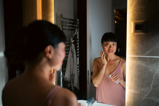 Asian woman applying skincare cream in bathroom mirror