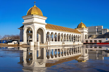 Naklejka premium Colonnaded pavilion with golden domes near the Romanian National Cathedral in Bucharest, reflected in rainwater under a clear blue sky in the capital of Romania.