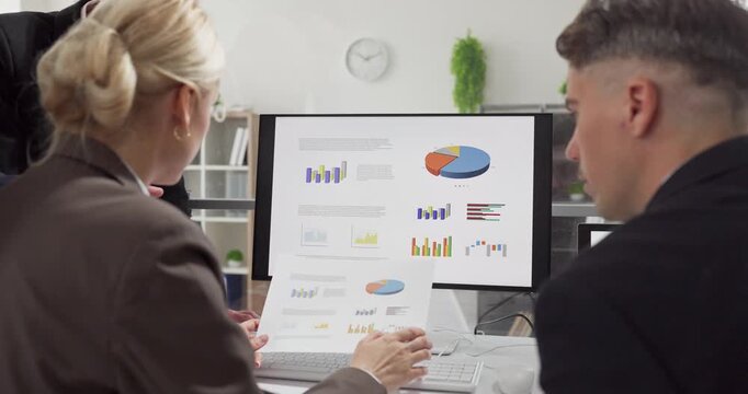 Team colleagues meeting in office computer charts. Analysts compare a report printout with data on the monitor with metrics for planning and discussion. Concept image of office teamwork.