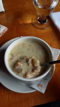 Bowl of traditional New England clam chowder with spoon on table. Close up of white ceramic bowl filled with creamy seafood soup containing potatoes and herbs on a wooden surface.