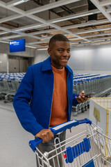 A young African man with short hair smiles while pushing a shopping cart in a modern store. He holds a smartphone in his hand, surrounded by shopping carts.