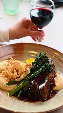 Braised beef rib served with red wine in restaurant. Meat dish with mashed potatoes and green vegetables paired with glass of red wine.
