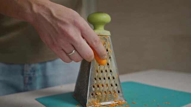 Close-up shot of hands grating a carrot on a box grater. Detailed vegetable preparation, food texture and everyday home cooking process. High quality 4k footage