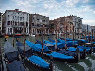 Moored Gondolas with Grand Canal Palaces - A row of traditional Venetian gondolas covered with blue tarps moored on the water, with historic Grand Canal palaces and buildings in the background.