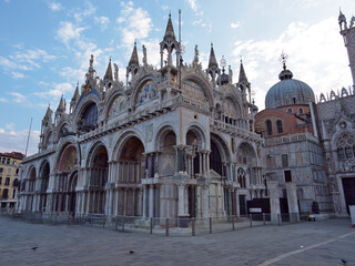 Fototapeta premium St Mark's Basilica Facade and Mosaics - Wide, majestic view of the ornate Byzantine facade of St. Mark's Basilica with marble arches, detailed mosaics, and spires under a cloudy blue sky.
