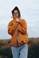 Young woman lifestyle in orange jacket and glasses stands outdoors in a field under cloudy sky. Casual fashion and autumn mood concept with natural background and thoughtful expression.