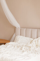 Unmade white bed with beige upholstered headboard and sheer canopy in minimalist bedroom, beige wall background, wooden nightstand. Cozy home, morning routine, hospitality, comfort.