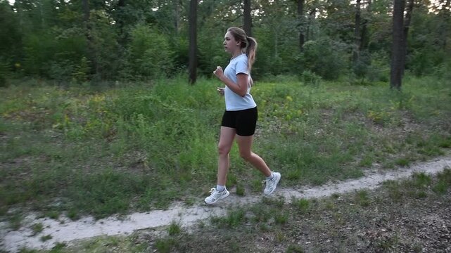 Young woman running along dirt park trail in slow motion with perpendicular camera movement. Side view while camera pulls away creates dynamic depth and cinematic perspective.