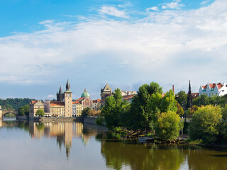 Fototapeta premium Vltava River Prague Panorama Sunny Day - Scenic view of the Vltava River, historical buildings, and green trees under a bright blue sky.