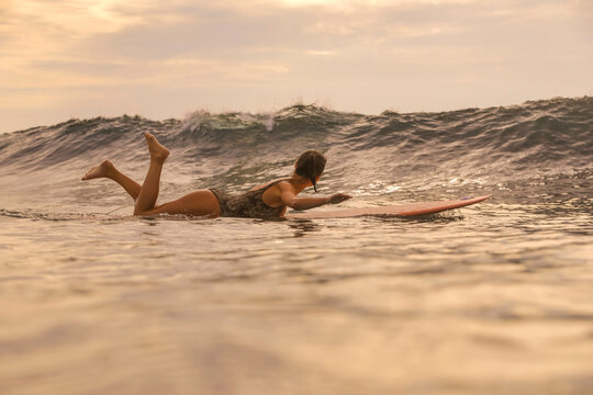 Young Woman Paddling On Surfboard At Sunset, Lying Prone And Stroking Toward Horizon, Warm Golden Light