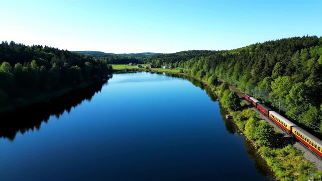 A train travels along a lake, surrounded by green trees on a sunny day. The clear blue sky complements the tranquil landscape