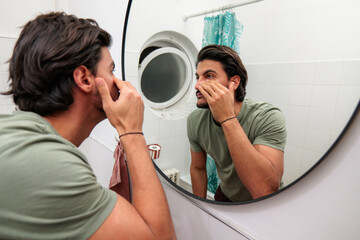 Young man examining his face in a bathroom mirror, practicing a skincare and grooming routine at home, focused on appearance, hygiene and everyday self care