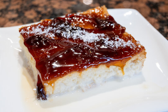 Close-up of a Trilece cake slice on a white plate. Sponge base soaked in milk, topped with glossy caramel and shredded coconut. A bite reveals the moist, layered composition. 