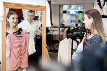In clothing store, girl holds hanger with blouse and looks at her reflection in combination with...