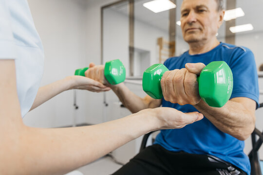 Senior man performing therapeutic exercises with green dumbbells while receiving guidance from a female therapist in a bright rehabilitation center
