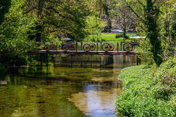 Obraz premium A decorative wooden footbridge, with antique wagon wheels, spans a shallow, sunlit forest stream surrounded by dense, rich green foliage near Saint Naum Monastery on Lake Ohrid, North Macedonia
