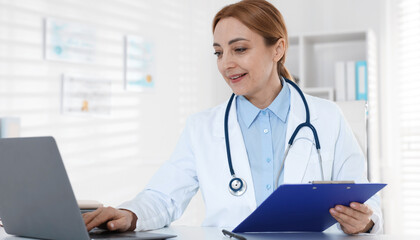 Smiling cardiologist with laptop and clipboard working at table in clinic