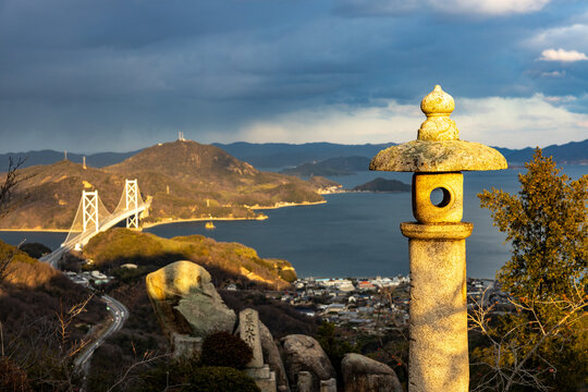 Stone lantern overlooking the Seto Inland Sea and Shimanami Kaido bridge in Hiroshima Prefecture, Japan. Scenic coastal landscape under dramatic evening sky.