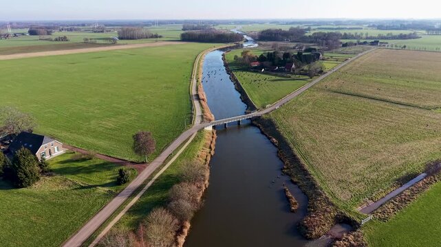 Aerial view of a rural landscape with a canal curving through green fields, two small road bridges crossing it, and a nearby house with outbuildings set among open farmland.