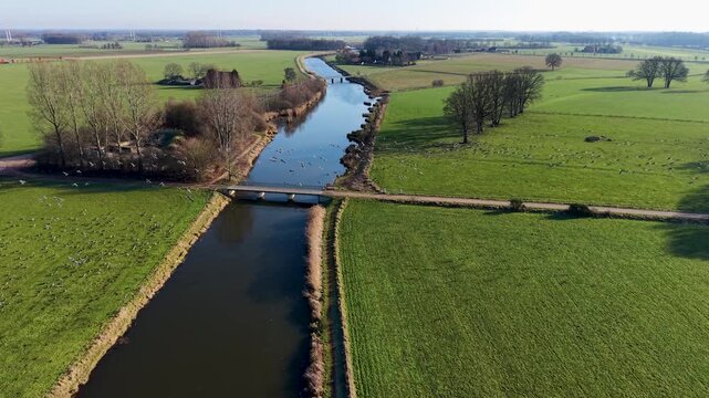 Aerial view of a rural landscape with a canal curving through green fields, two small road bridges crossing it, and a nearby house with outbuildings set among open farmland.