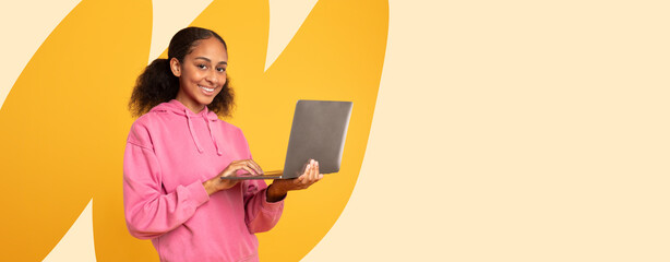 A young person stands holding a laptop with a smile on their face. The background features a simple design with a bright shade. It is a moment of engagement with technology. © Prostock-studio
