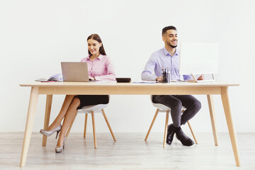 Two individuals are focused on their tasks at a shared office desk. A woman uses a laptop while a...