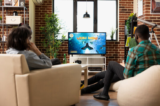 Male and female friends sit around television displaying you win message during gaming hangout at home. Young group celebrates winning online video game together as team in apartment.