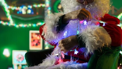 Fototapeta premium Professional Santa Claus knotted in xmas lights while decorating room with stockings, fireplace, ornaments and other holiday props, trying to get untangled from Christmas led lights, camera A