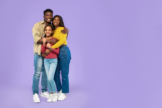 A father, mother, and daughter pose closely, smiling while embracing each other. The family shows happiness and unity while standing against a solid purple background in a studio setting.