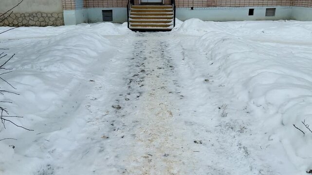 A snow-covered unmade path to the entrance to the pharmacy, a narrow trampled path through deep snow with sand and slush, leading iron steps.